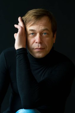 45-50 yaşlarında, siyah arka planda golf kazağı giyen, kameraya bakan ciddi bir adamın portresi. Belki de eski bir asker, iş adamı, avukat ya da karizmatik bir aktördür..