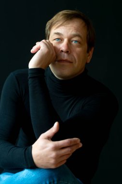 45-50 yaşlarında, siyah arka planda golf kazağı giyen, kameraya bakan ciddi bir adamın portresi. Belki de eski bir asker, iş adamı, avukat ya da karizmatik bir aktördür..