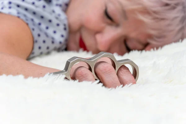 A metal brass knuckles in the hand of an elderly woman 60-65 years old sleeping in bed. Concept: domestic violence, self-defense in the apartment, family quarrels.