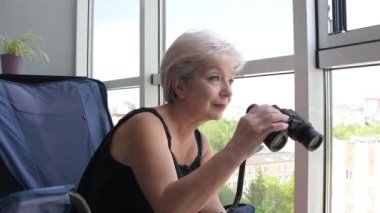 60-65 yaşlarındaki yaşlı bir kadın karşıdaki evden dürbünle gözetliyor. Konsept: komşuları gözetleme, bilgi toplama, casusluk ve dedikodu.