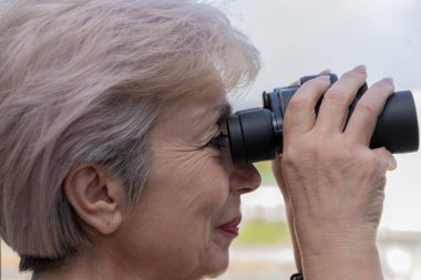 60-65 yaşlarındaki yaşlı bir kadın karşıdaki evden dürbünle gözetliyor. Konsept: komşuları gözetleme, bilgi toplama, casusluk ve dedikodu.