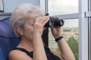 60-65 yaşlarındaki yaşlı bir kadın karşıdaki evden dürbünle gözetliyor. Konsept: komşuları gözetleme, bilgi toplama, casusluk ve dedikodu.