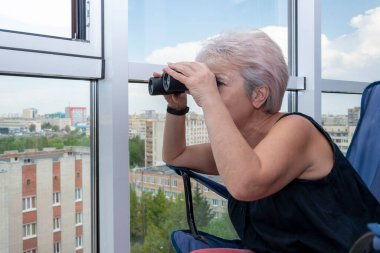 60-65 yaşlarındaki yaşlı bir kadın karşıdaki evden dürbünle gözetliyor. Konsept: komşuları gözetleme, bilgi toplama, casusluk ve dedikodu.