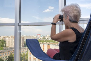 60-65 yaşlarındaki yaşlı bir kadın karşıdaki evden dürbünle gözetliyor. Konsept: komşuları gözetleme, bilgi toplama, casusluk ve dedikodu.