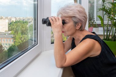60-65 yaşlarındaki yaşlı bir kadın karşıdaki evden dürbünle gözetliyor. Konsept: komşuları gözetleme, bilgi toplama, casusluk ve dedikodu.
