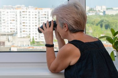 60-65 yaşlarındaki yaşlı bir kadın karşıdaki evden dürbünle gözetliyor. Konsept: komşuları gözetleme, bilgi toplama, casusluk ve dedikodu.