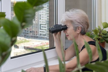 An elderly woman of 60-65 years old peeps through binoculars at neighbors from the house opposite. Concept: spying on neighbors, gathering information, espionage and gossip.