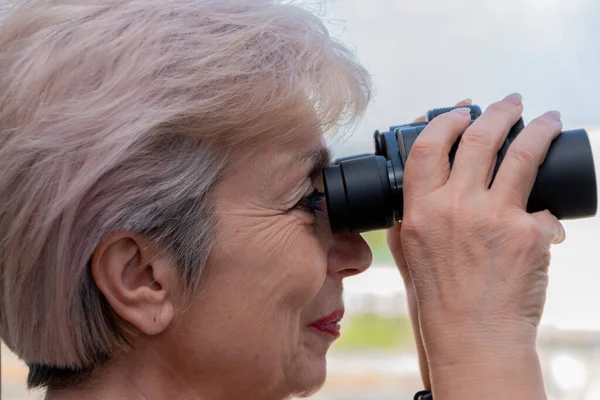 60-65 yaşlarındaki yaşlı bir kadın karşıdaki evden dürbünle gözetliyor. Konsept: komşuları gözetleme, bilgi toplama, casusluk ve dedikodu.