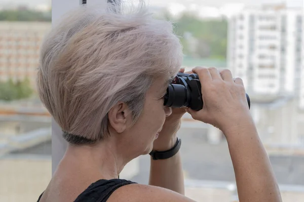 An elderly woman of 60-65 years old peeps through binoculars at neighbors from the house opposite. Concept: spying on neighbors, gathering information, espionage and gossip.