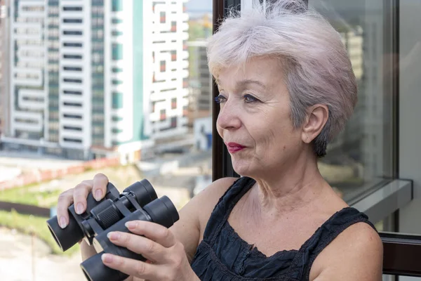 An elderly woman of 60-65 years old peeps through binoculars at neighbors from the house opposite. Concept: spying on neighbors, gathering information, espionage and gossip.