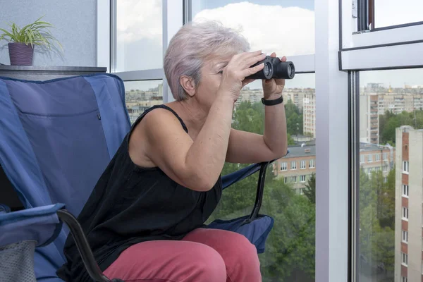 60-65 yaşlarındaki yaşlı bir kadın karşıdaki evden dürbünle gözetliyor. Konsept: komşuları gözetleme, bilgi toplama, casusluk ve dedikodu.