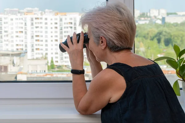 60-65 yaşlarındaki yaşlı bir kadın karşıdaki evden dürbünle gözetliyor. Konsept: komşuları gözetleme, bilgi toplama, casusluk ve dedikodu.