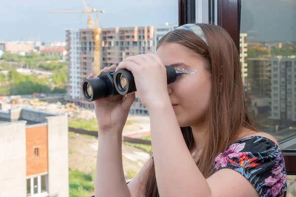 A young girl from the window is watching the street and neighboring houses through binoculars. Concept: spying on neighbors, gathering information, espionage and gossip, looking out for the groom.