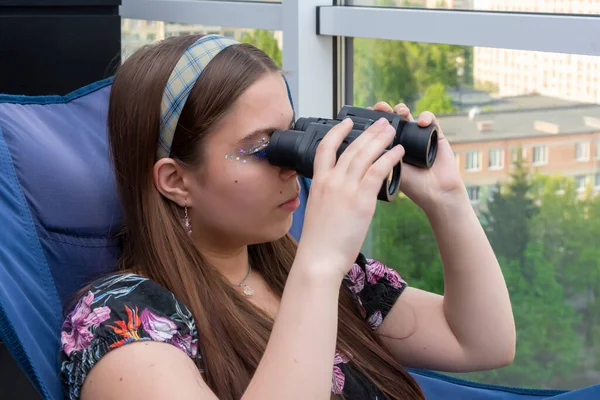 18-20 yaşlarında bir kız dairesinin penceresinden sokakta dürbünle bakıyor. Kavram: komşuları gözetlemek, bilgi toplamak.