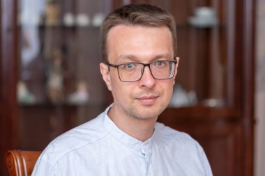 Gözlüklü orta yaşlı bir adam ve evin arka planında bulanık duran açık renk bir gömlek..