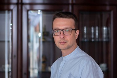 Gözlüklü orta yaşlı bir adam ve evin arka planında bulanık duran açık renk bir gömlek..