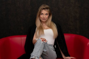 20-25 yaşlarında, güzel, seksi bir sarışın. Uzun saçlı. Kırmızı koltukta oturuyor..