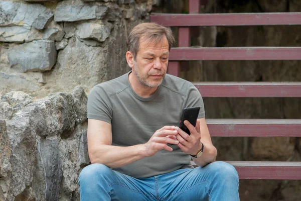 Street Portrait of an elderly man 45-50 years old in a T-shirt in summer, taking a selfie on his phone against the background of an old stone wall.