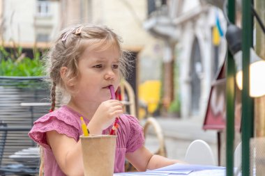 Street portrait of a beautiful 4-year-old girl drawing with colored pencils at a table in an outdoor cafe on a blurry background of the old town.
