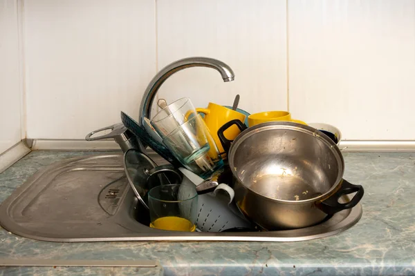 Dirty and unwashed dishes are stacked in the kitchen sink. Unwashed cups, plates, pots, forks and spoons.The height of the water tap determines the number of dishes in the sink.