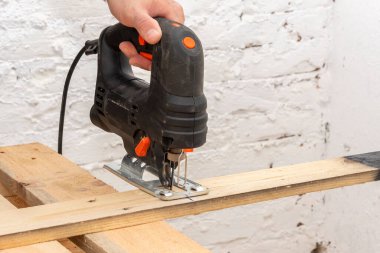 The carpenter's hand holds an electric jigsaw and cuts a wooden board, close-up. Carpentry in the workshop.