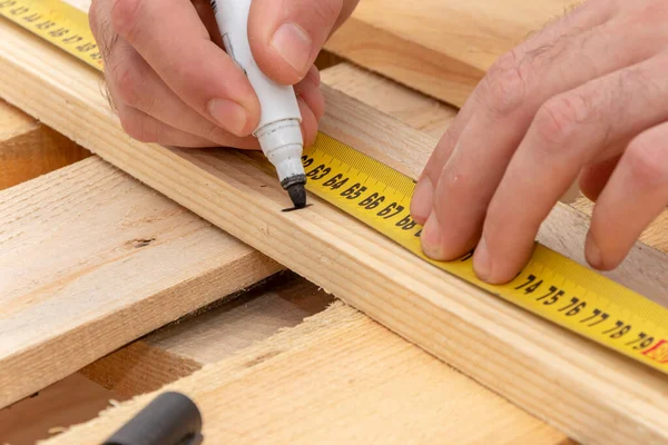 The carpenter puts a marker marker, measures a wooden board with a tape measure, close-up.