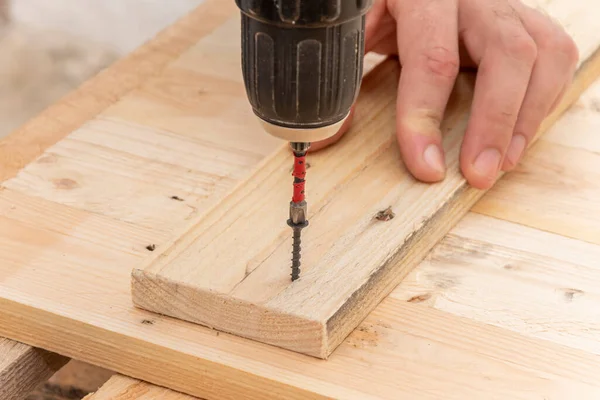 Close-up details of a Caucasian male carpenter using an electric screwdriver in his hand and repairing a new wooden table, home improvement concept