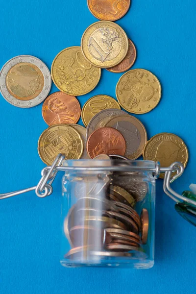 Small coins are poured out of a glass jar on a blue background, top view. Concept: financial crisis, decrease in income, capital investments.