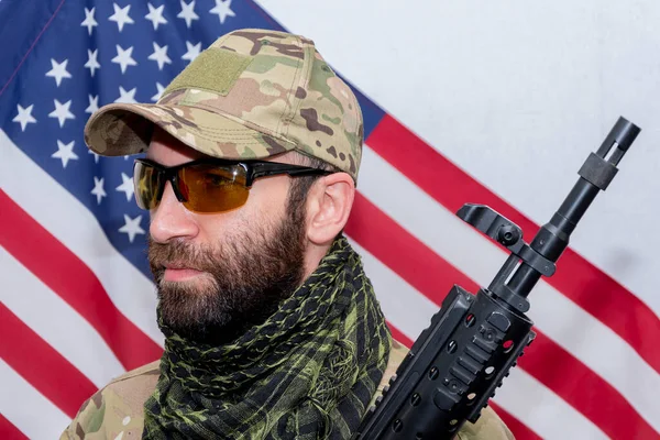 Portrait of a military man 30-35 years old with a rifle on his shoulder, wearing dark glasses against the background of the American flag.Concept: military action, soldier of fortune, US Army.