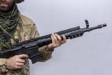 A military man in camouflage holds an American automatic rifle in his hands on a light background, close-up. Concept: military action, soldier of fortune.
