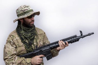 A military man in camouflage and a panama hat holds an American automatic rifle in his hands on a light background, close-up. Concept: military action, soldier of fortune.