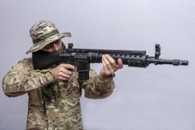 A military man in camouflage and a panama hat aims an American rifle on a light background, close-up. Concept: military action, soldier of fortune.