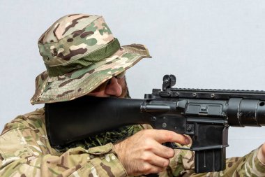 A military man in camouflage holds an American automatic rifle in his hands on a light background, close-up. Concept: military action, soldier of fortune.