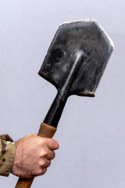 A soldier holds a sharpened sapper shovel in his hand on a light background.