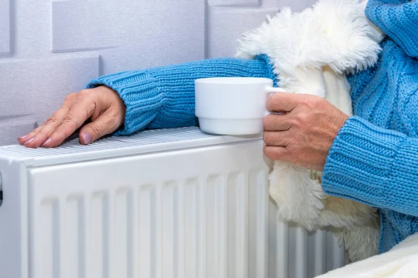 Elderly woman warming her hands on the heating radiator, holding a cup in her hand. Concept: low room temperature, rising gas and heating prices, coldness in the apartment.