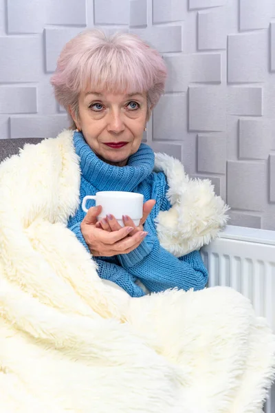 Portrait of an elderly woman 60-65 years old, holding a white cup in her hands against a heating radiator. Concept: low room temperature, rising rent, coldness in the apartment.