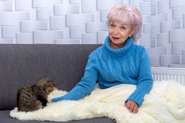 Portrait of a beautiful elderly woman of 60-65 years, sitting on the couch with a kitten, covered with a warm plaid. Concept: a cold snap in the apartment, rising electricity and gas prices.