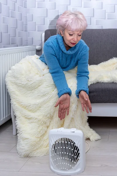 An elderly woman of 60-65 warms her hands from the heater, sitting on the couch, covered with a warm plaid. Concept: a cold snap in the apartment, rising electricity and gas prices.