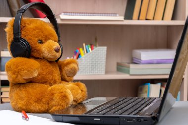 Bilgisayarında kulaklıkları olan oyuncak ayı kitapların ve belgelerin arka planında oturuyor. Konsept: uzaktan çalışma, internet üzerinden iletişim, evde çalışma.