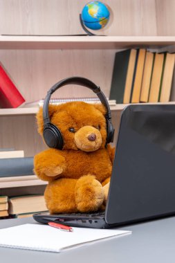Kulaklık takmış oyuncak ayı dizüstü bilgisayarın başında kitap raflarının arkasında oturuyor. Konsept: Uzak çalışma, internet üzerinden iletişim.