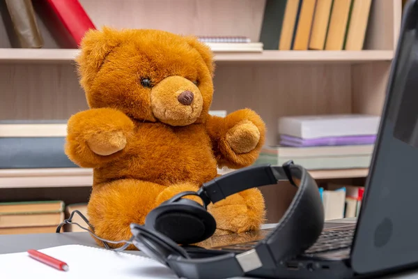 Toy teddy bear sitting on the table, next to the laptop and headphones against a background of shelves of books. The concept: remote work, communication via the Internet, work at home.
