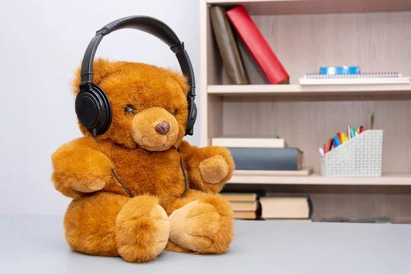 A teddy bear wearing headphones sits on a table against a neutral background in a room. The concept: communication via the Internet, remote press conference, listening to music.