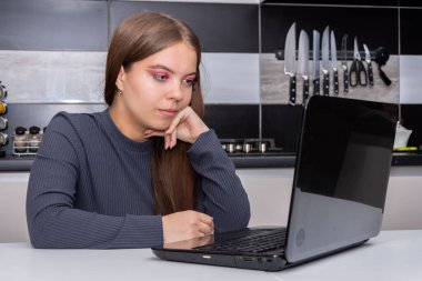 Genç bir kız, bulanık bir mutfak arka planında dizüstü bilgisayarının önünde dalgın dalgın oturuyor. Konsept: Coronavirüs zamanında uzaktan öğrenme.