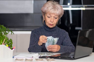 60-65 yaşlarında yaşlı bir kadın masasında dizüstü bilgisayarının önünde oturmuş para sayıyor. Konsept: fiyat artışları, elektrik faturaları, emeklilik.