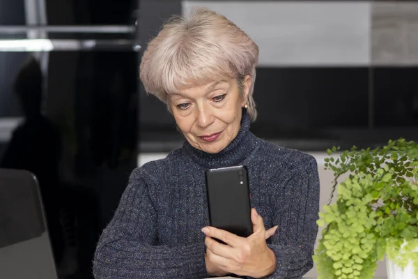 An elderly, beautiful woman looks at her smartphone against a neutral background. Concept: communication via the Internet, calling relatives.