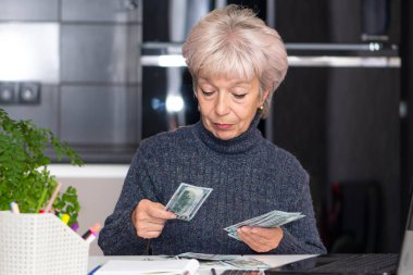 60-65 yaşlarındaki yaşlı bir kadın bilgisayarının önünde masasında oturan 100 dolarlık banknotları sayıyor. Kavram: daha yüksek fiyatlar, küçük emekli aylıkları, daha yüksek enerji ve ısıtma maliyetleri.