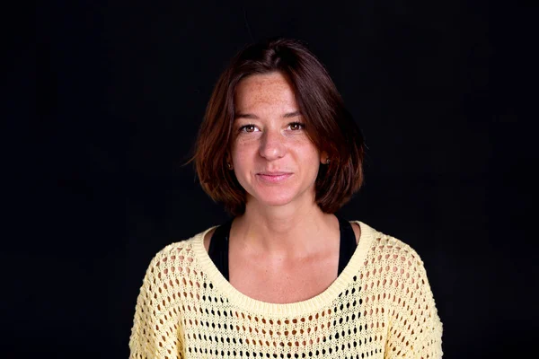 Studio portrait of a pretty woman of 35-40 with a freckled face against a dark background.