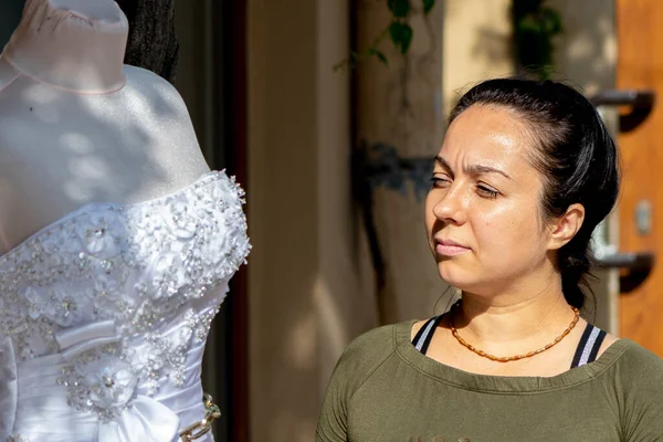 An adult, beautiful woman of 35-40 is looking at a wedding dress on a street mannequin. Concept: preparation for the wedding, desire and dream of getting married.