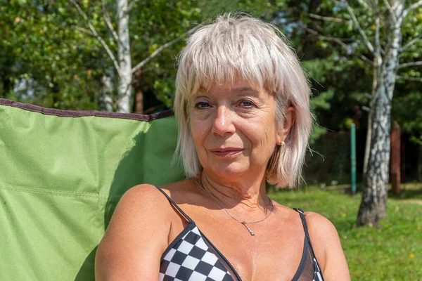 Portrait of a beautiful blonde woman 60-65 years old sits in a tourist chair, in a swimsuit against the backdrop of nature.