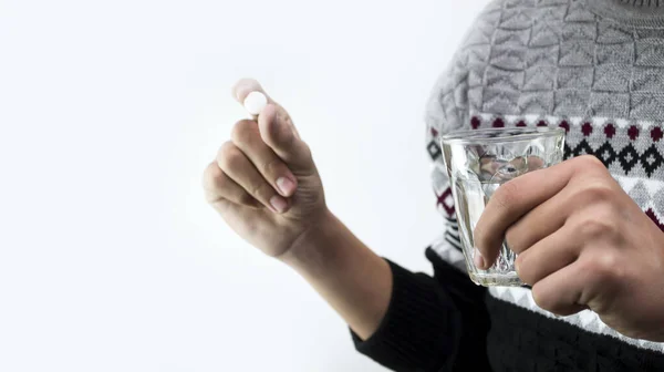 Imagem de um jovem segurando um copo de água em uma mão e um comprimido branco na outra. O homem está vestido com uma camisola cinzenta. o medicamento — Fotografia de Stock